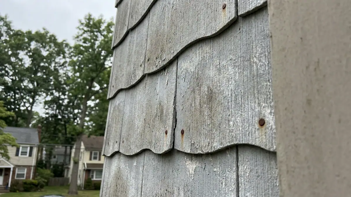 Close-up of weathered gray cement shingle siding with overlapping wavy-bottom panels, a chalky surface, one cracked shingle and old nail heads in muted daylight