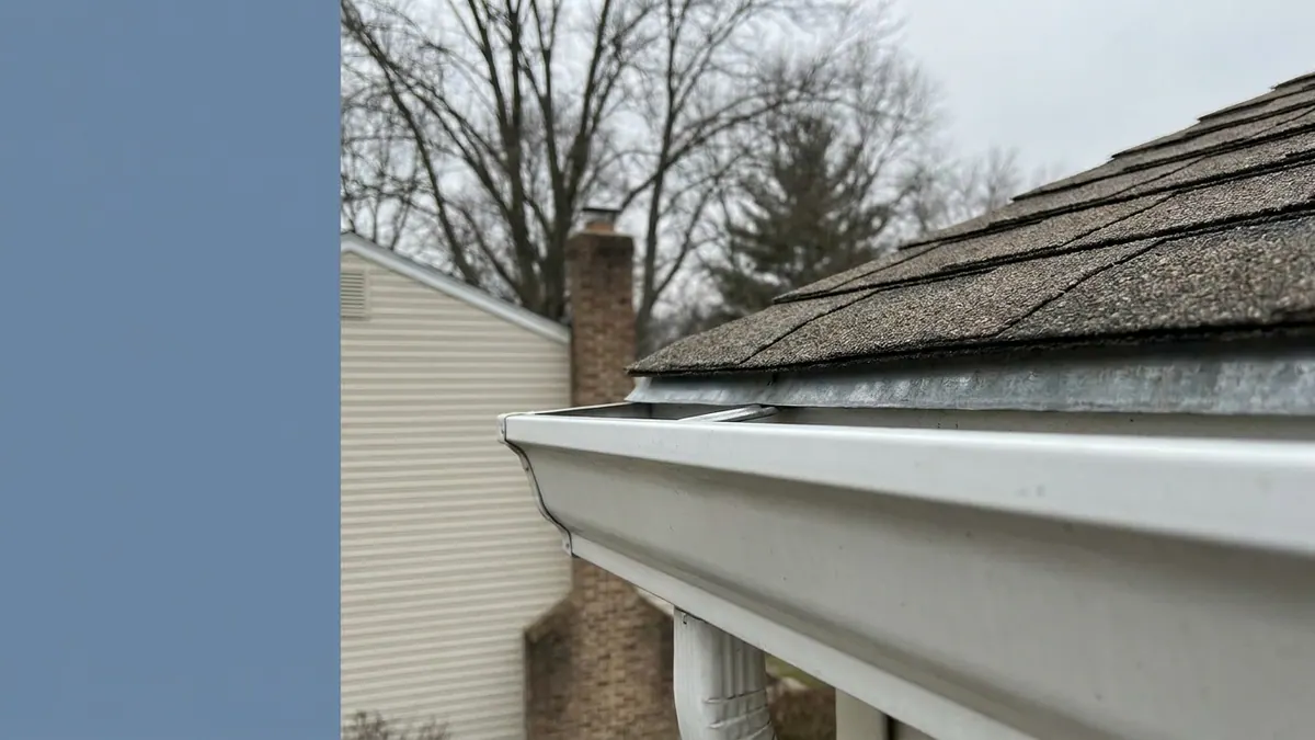 Close-up of a roof eave where the asphalt shingle edge meets a white aluminum K-style gutter, with the metal edge flashing tucked behind the gutter lip under overcast light