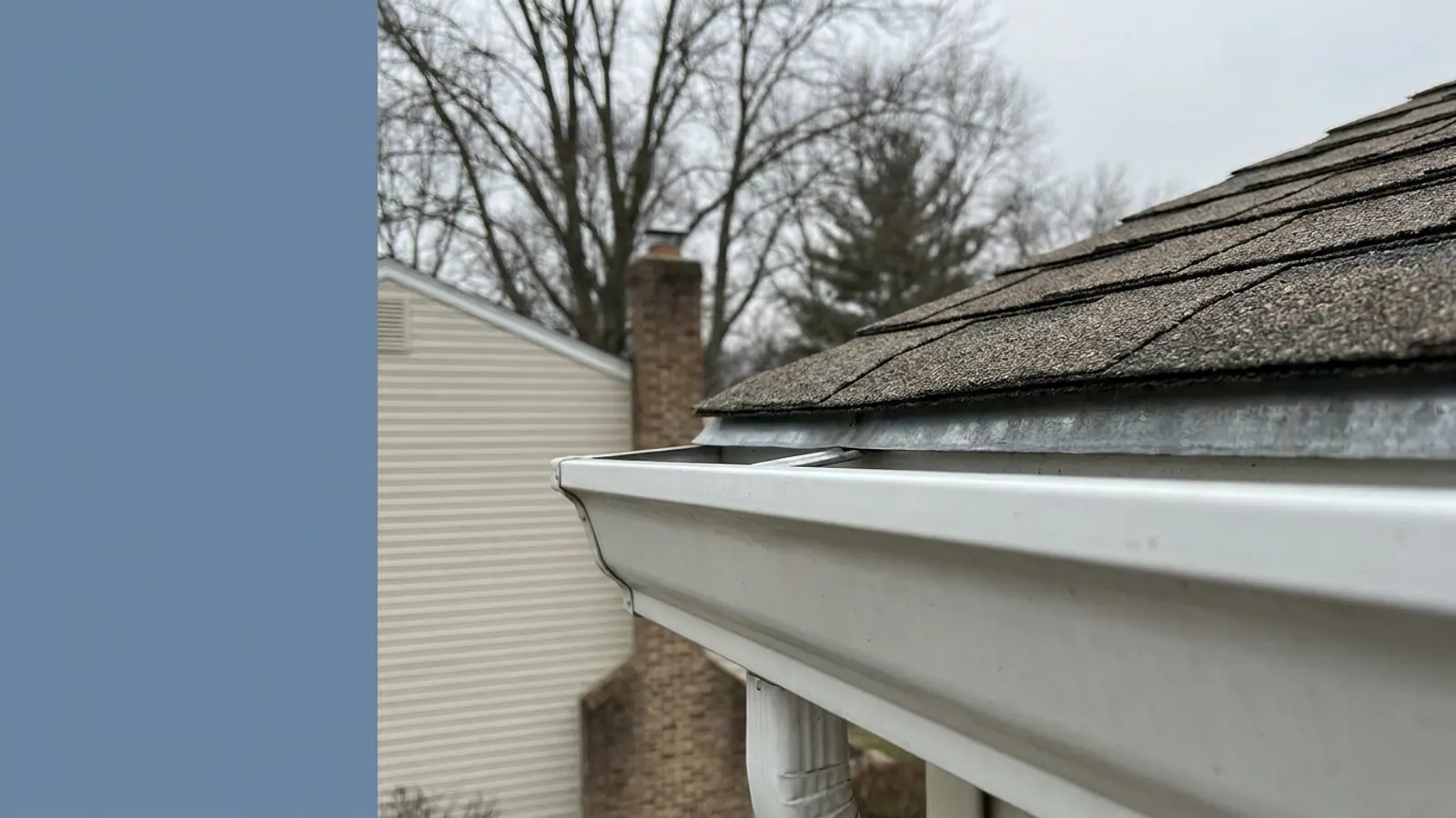 Close-up of a roof eave where the asphalt shingle edge meets a white aluminum K-style gutter, with the metal edge flashing tucked behind the gutter lip under overcast light
