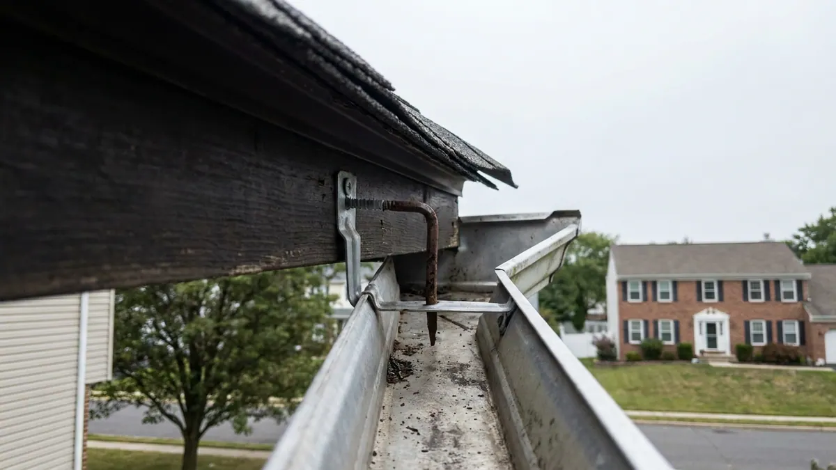 Close-up looking down into an open aluminum gutter trough along a fascia board, with a hidden hanger clip screwed into the fascia and an older rusted spike beside it, shingle roof edge above in morning light