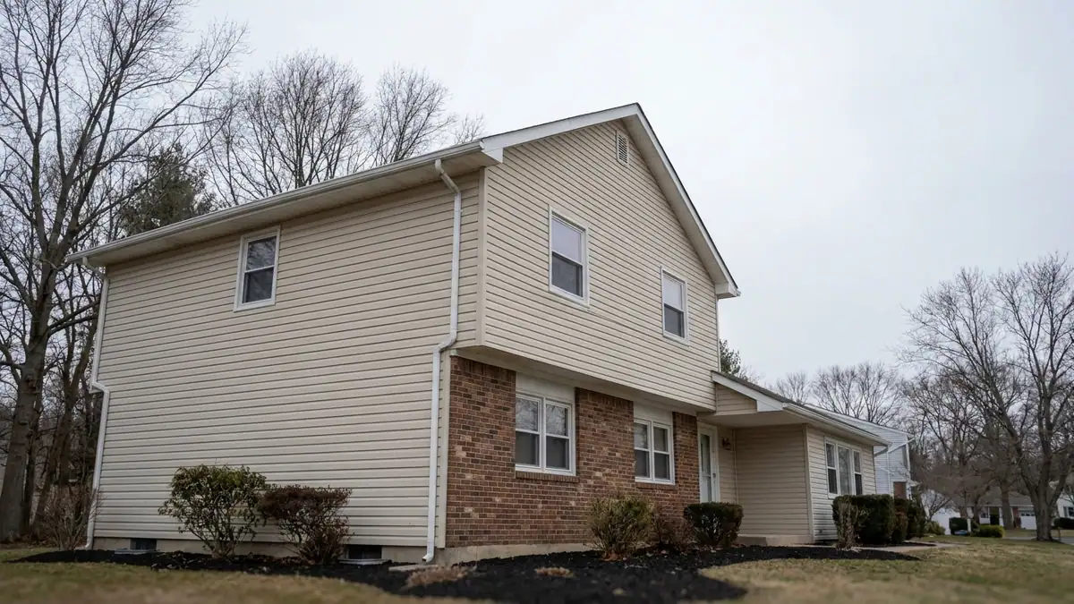 White seamless aluminum gutter running along one eave of a suburban split-level home beside a gable end with no gutter above mulch beds, soft cloudy daylight