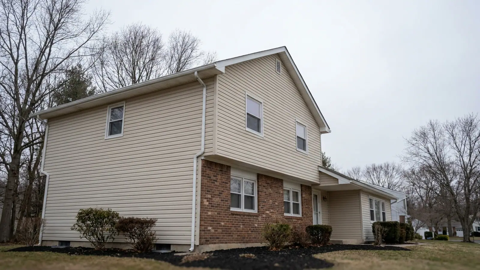 White seamless aluminum gutter running along one eave of a suburban split-level home beside a gable end with no gutter above mulch beds, soft cloudy daylight