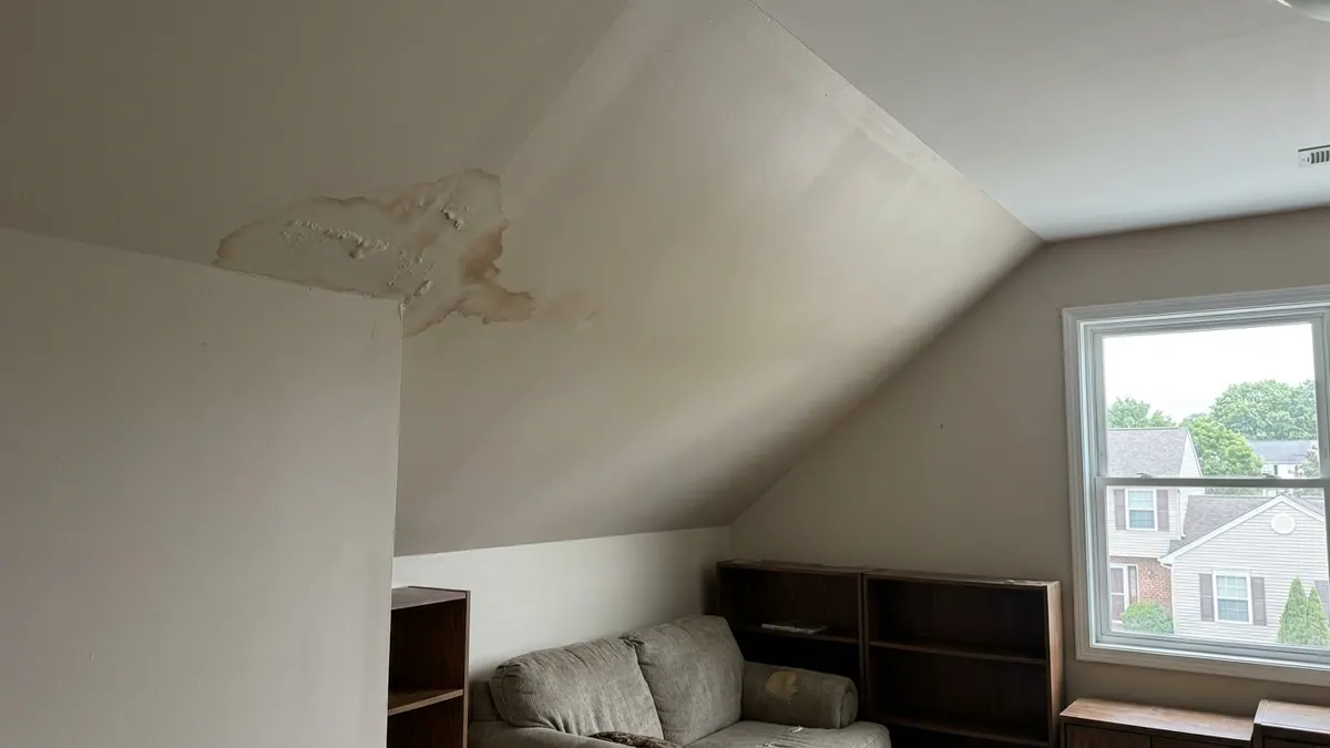 Sloped interior ceiling of a finished room above a garage with a brown water stain and bubbling paint near the joint where the ceiling meets the wall, lit by daylight from a side window