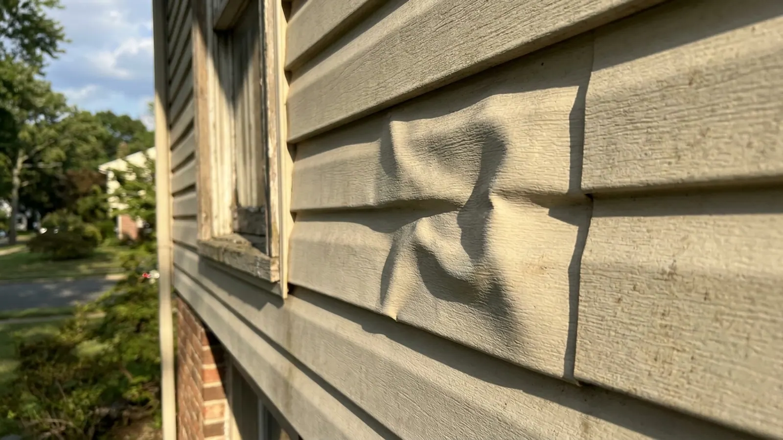 Close-up of light beige vinyl siding on a sunlit wall with one rippled, distorted panel course, sharp shadow lines showing the deformation next to flat undamaged siding