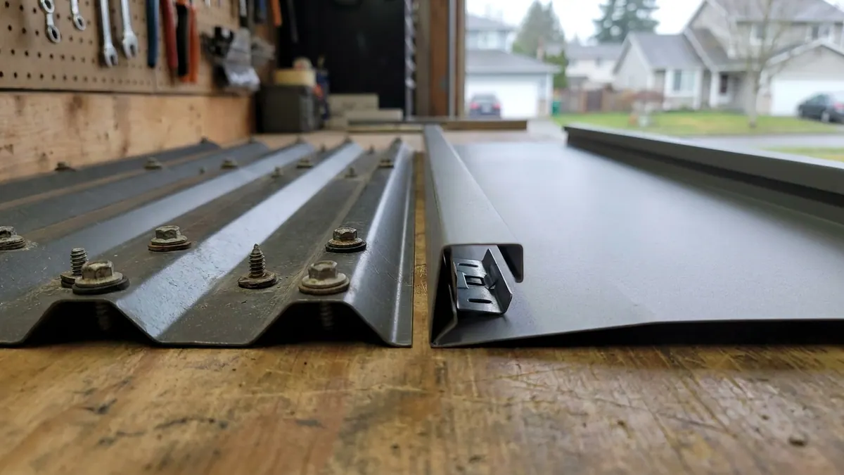 Two metal roofing panels side by side on a workbench in daylight, one ribbed panel with rows of gasketed screws through its face and one smooth standing-seam panel with a hidden clip resting beside it