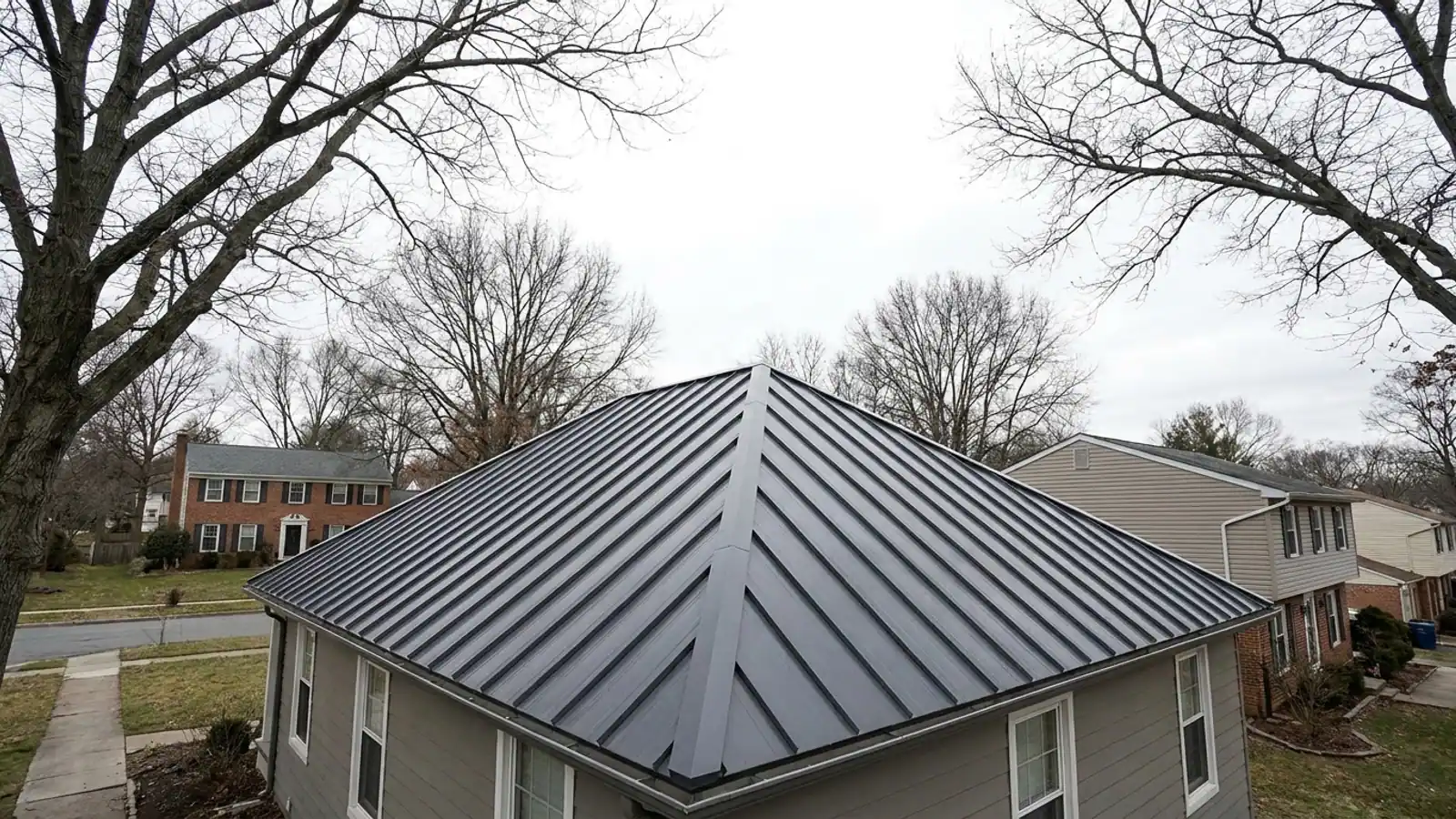 Ground-level view of a house with a dark standing seam metal hip roof, four sloping planes meeting at crisp ridge and hip lines with angled panel cuts along the hips, trees at the edge of frame