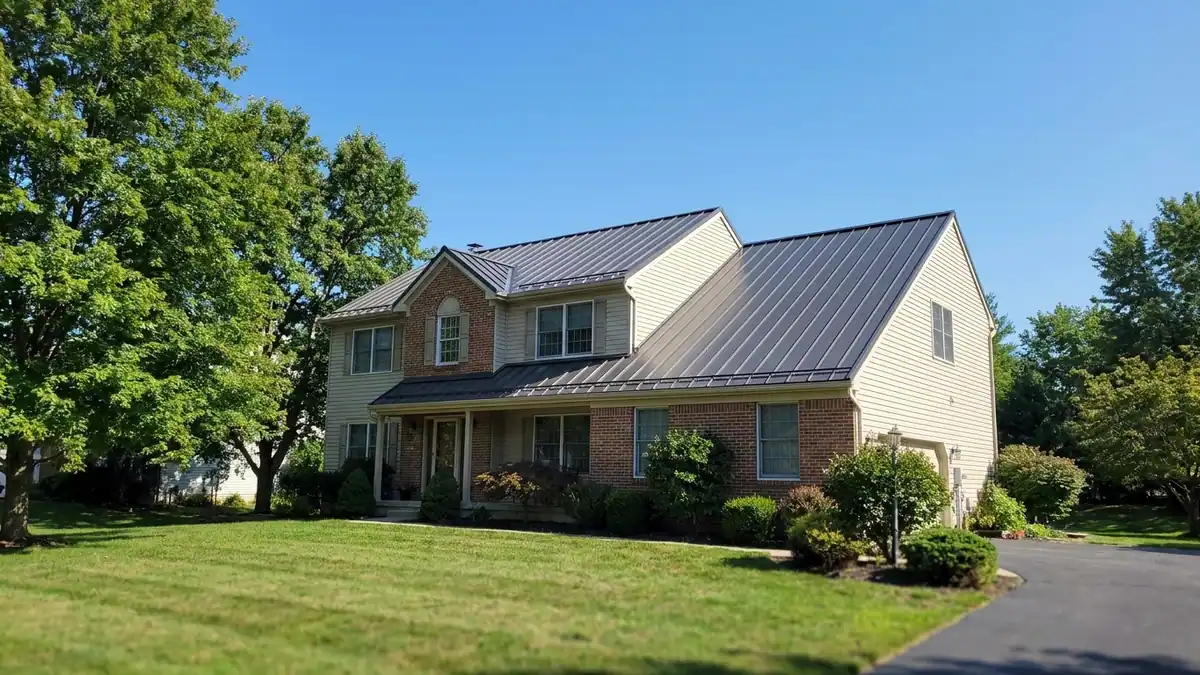 Dark standing seam metal roof on a two-story home seen from the front yard, crisp panel ridges and clean eaves visible under a clear sky with mature trees at one side