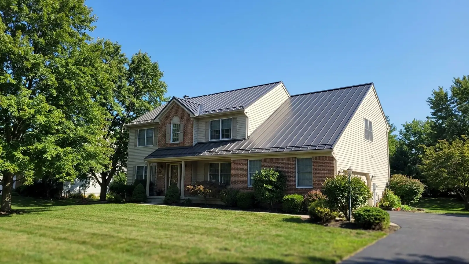Dark standing seam metal roof on a two-story home seen from the front yard, crisp panel ridges and clean eaves visible under a clear sky with mature trees at one side