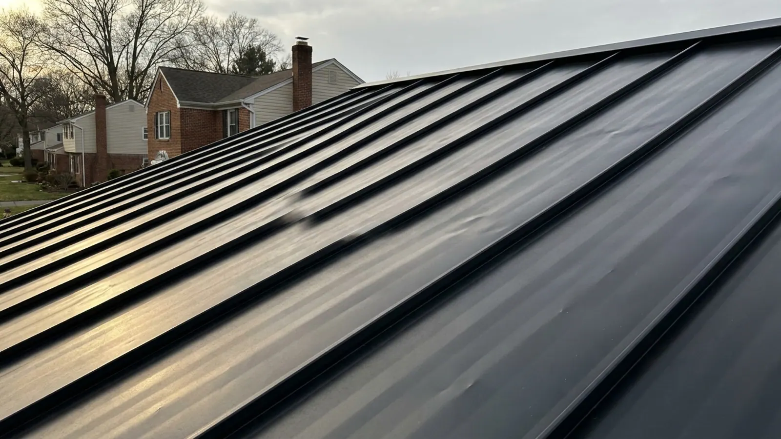 Close-up of a dark charcoal standing seam metal roof slope in low sunlight showing subtle waviness across the flat pan between sharp raised seams