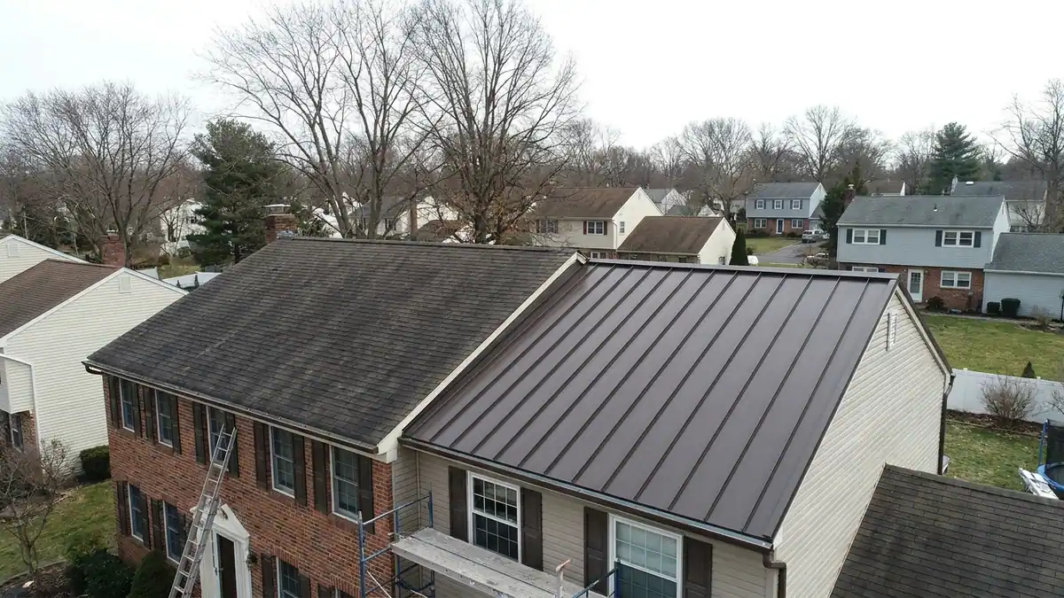 Residential roof mid-transition with dark asphalt shingles on one plane and new dark bronze standing-seam metal panels on the adjacent plane under a clear blue sky