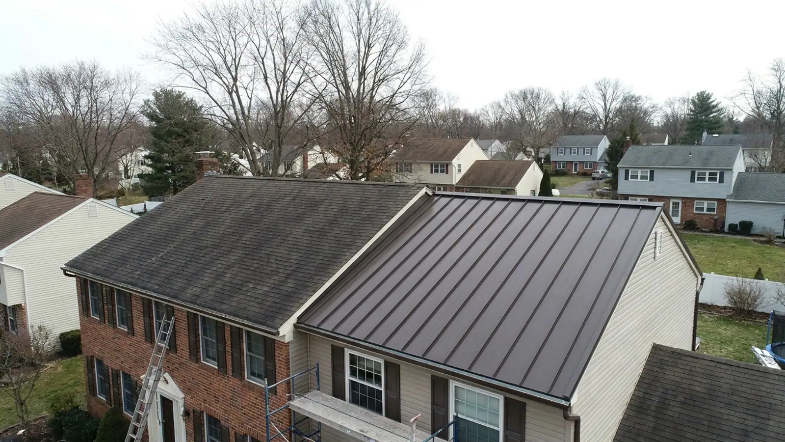 Residential roof mid-transition with dark asphalt shingles on one plane and new dark bronze standing-seam metal panels on the adjacent plane under a clear blue sky