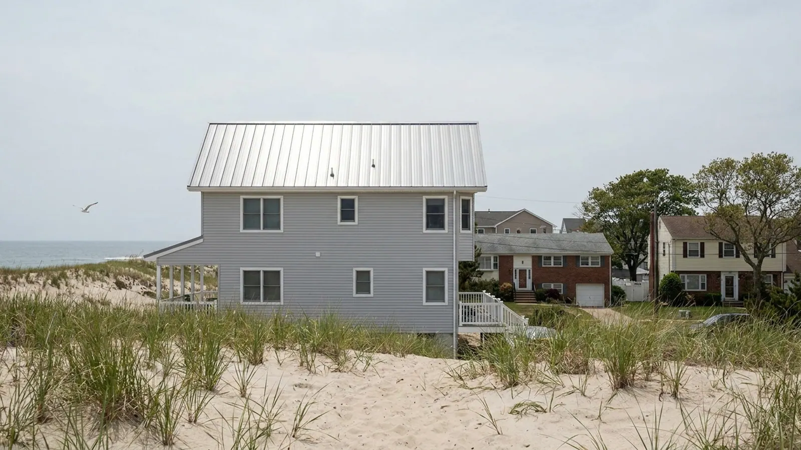 Light gray standing seam metal roof on a coastal house with dune grass in the foreground and bright hazy sky above