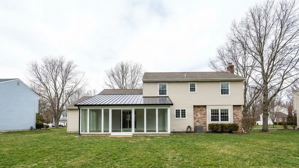 Sunroom addition with large glass walls and a low-slope standing seam metal roof attached to the rear of a suburban house, green lawn and clear daylight