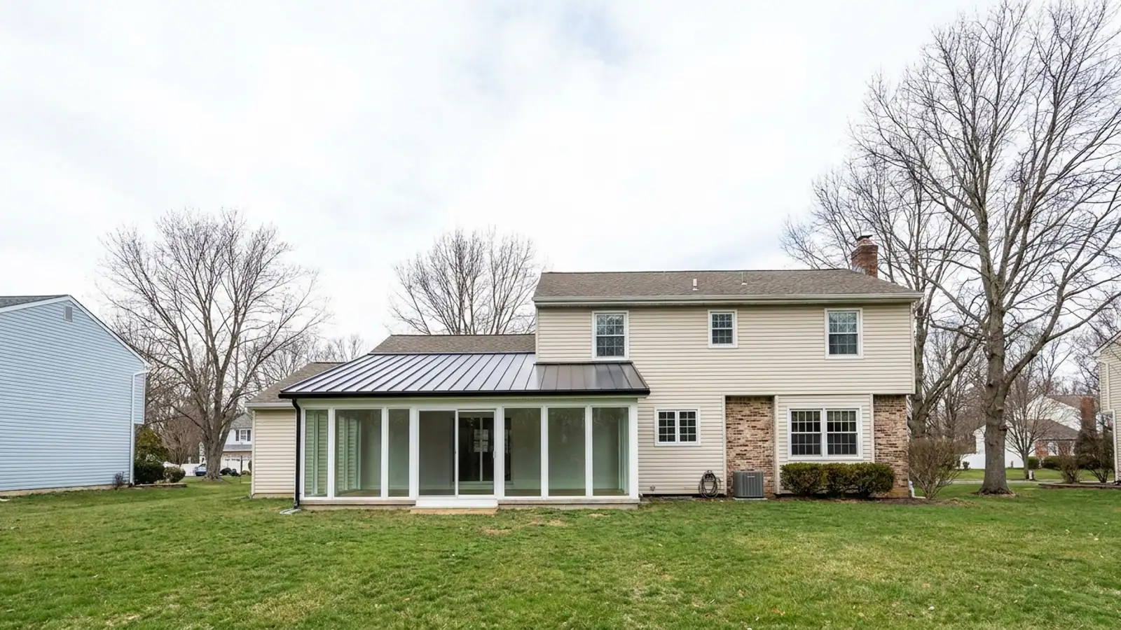 Sunroom addition with large glass walls and a low-slope standing seam metal roof attached to the rear of a suburban house, green lawn and clear daylight