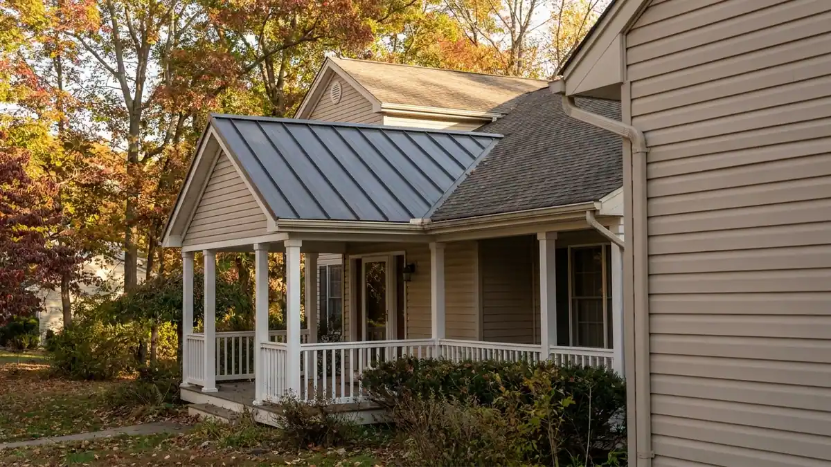 Low-slope standing-seam metal porch roof meeting a shingled main roof above, white porch columns and a painted railing in afternoon light