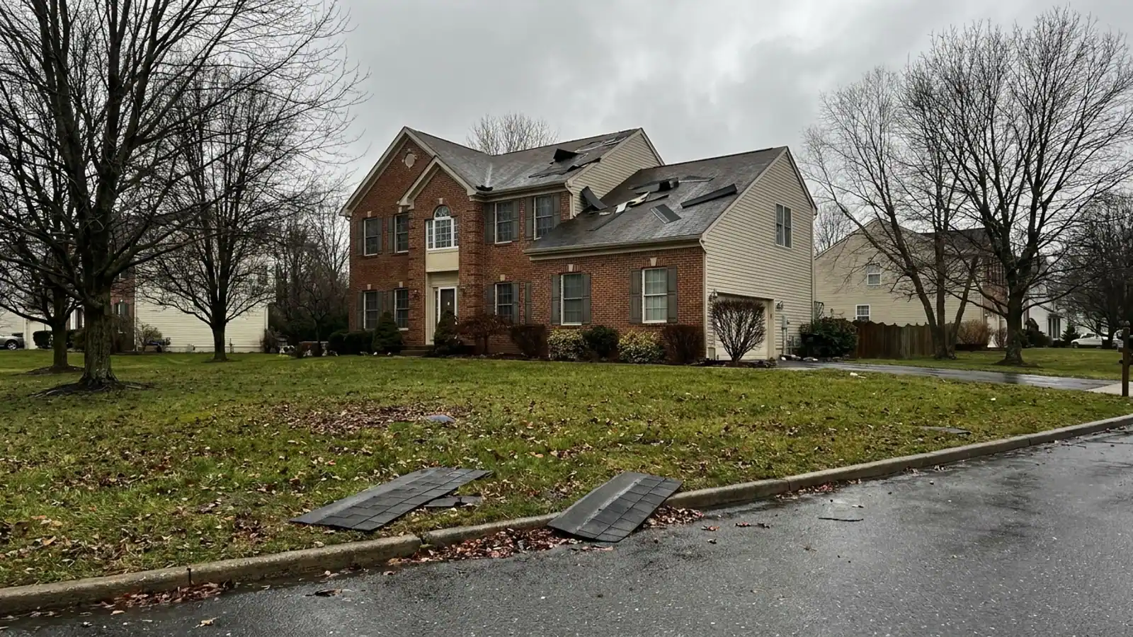 Asphalt shingles torn loose on a suburban roof slope after a storm with two loose shingles lying on wet grass in the foreground under an overcast gray sky