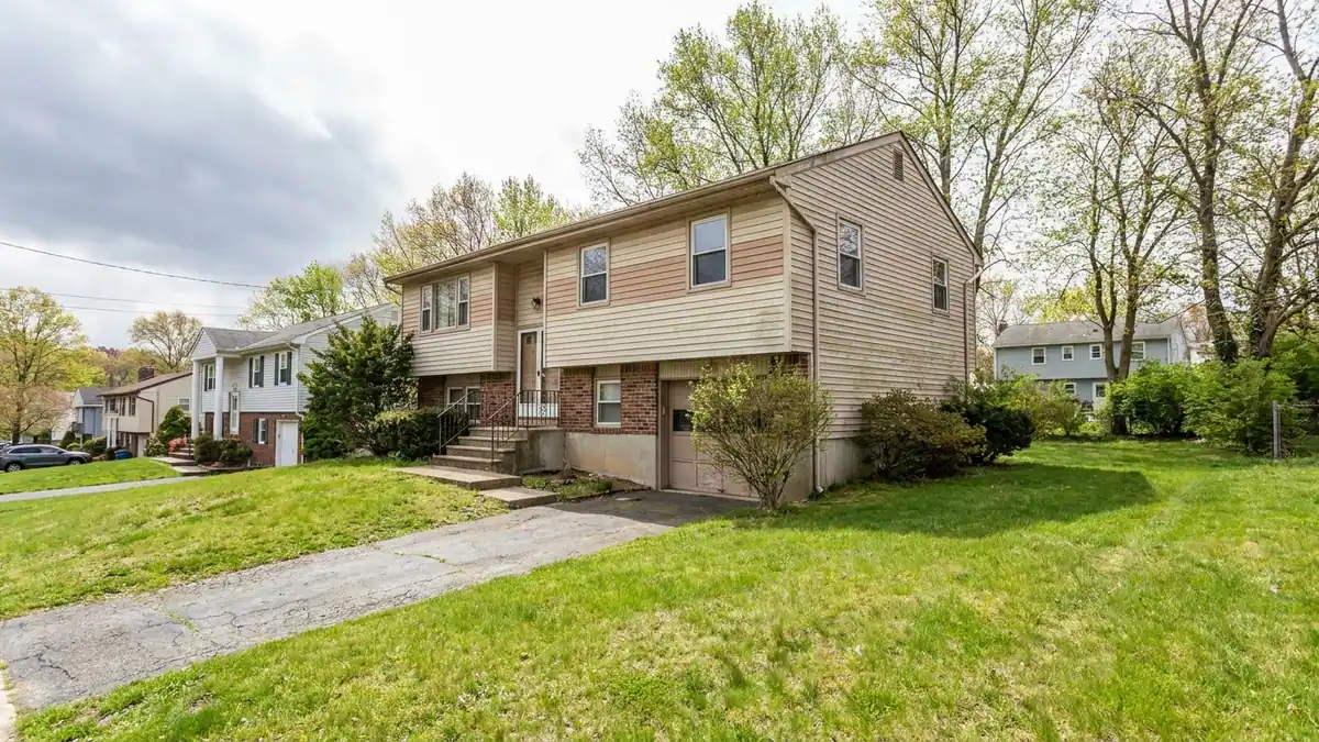 Raised ranch home viewed from the front corner with aged two-tone vinyl siding, a split entry with short front steps and a sloped driveway running down to the lower-level garage