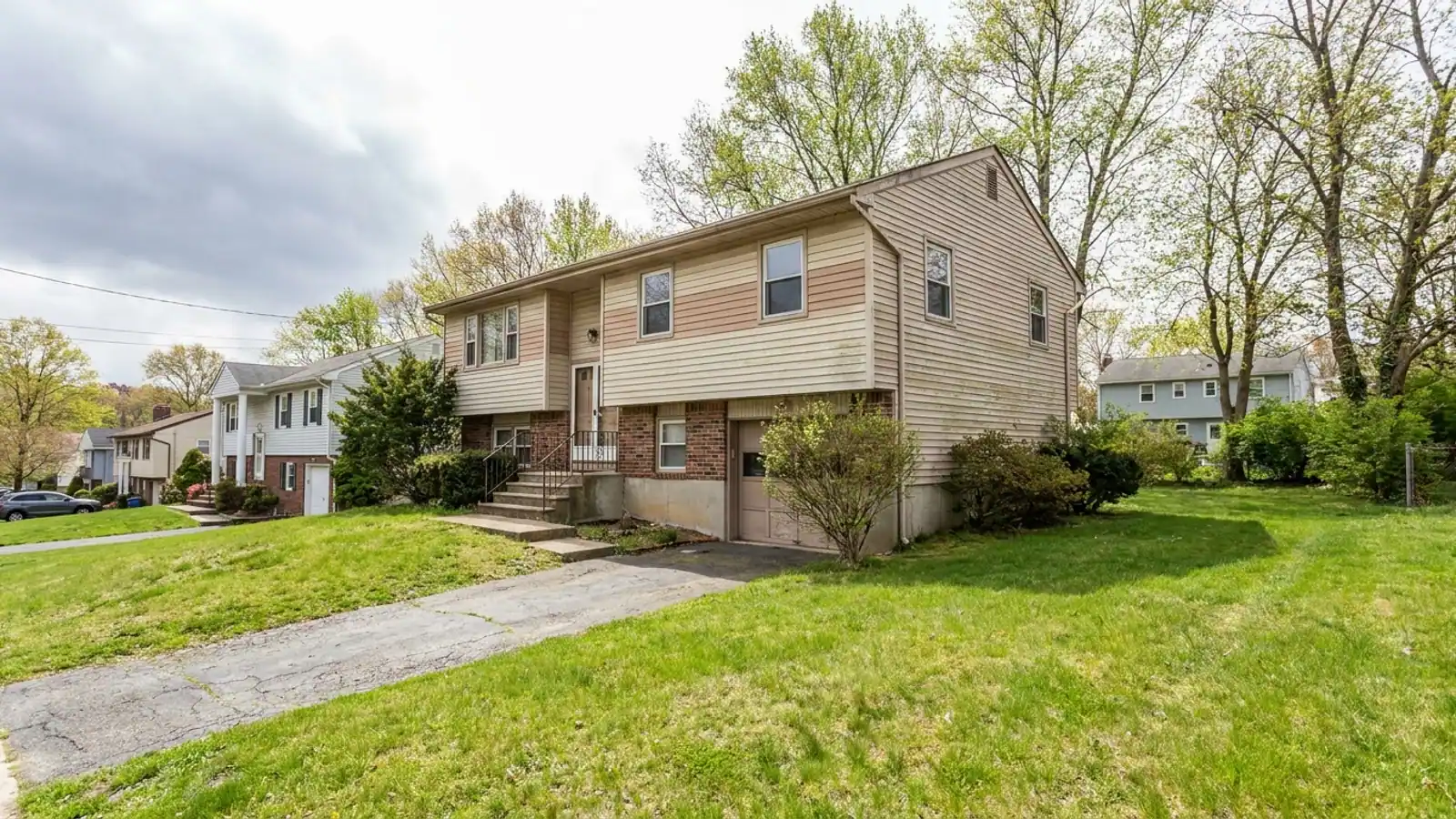Raised ranch home viewed from the front corner with aged two-tone vinyl siding, a split entry with short front steps and a sloped driveway running down to the lower-level garage