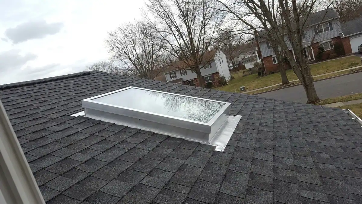 Curb-mounted skylight set into a dark asphalt shingle roof, aluminum flashing wrapped around the frame, clear sky reflected in the glass