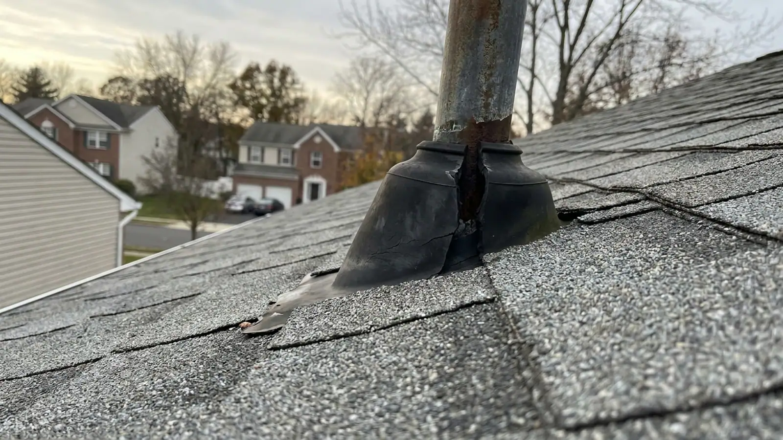 Close-up of a weathered rubber plumbing vent boot on an asphalt shingle roof with the collar cracked and split around the metal pipe under overcast light