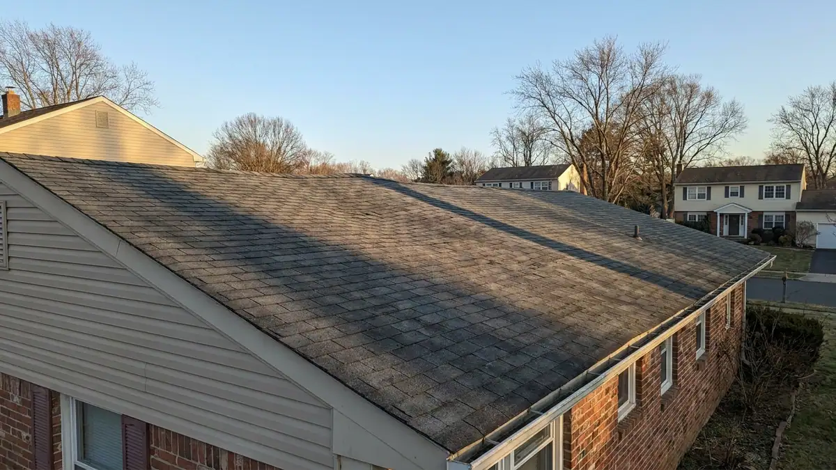 Low-angle view along the roof slope of an older single-story house in late afternoon light, showing a visible dip and wave in the shingle surface between rafters