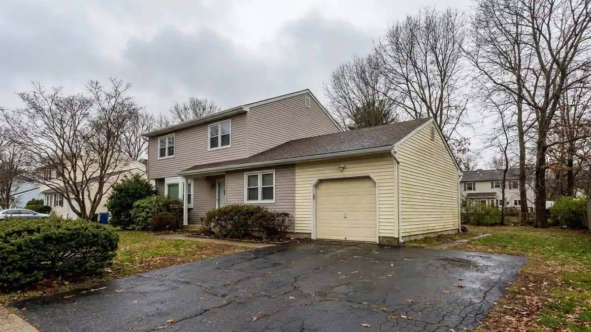 Two-story house with an attached garage wing clad in visibly faded vinyl siding a different shade from the main walls, a straight vertical seam where the two meet, driveway in the foreground under an overcast sky