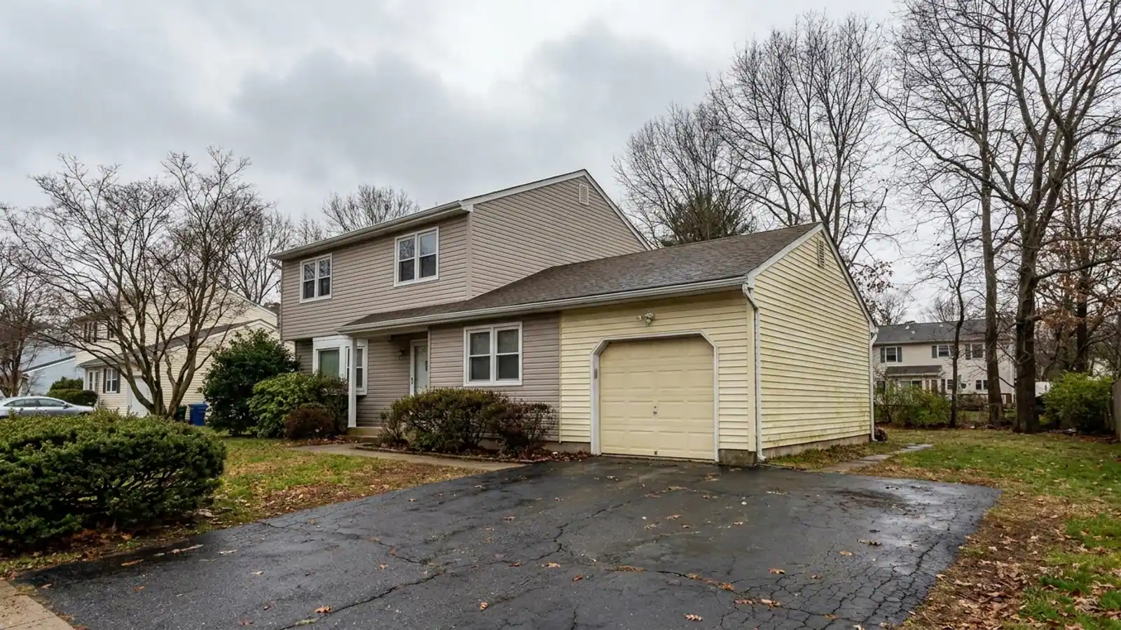 Two-story house with an attached garage wing clad in visibly faded vinyl siding a different shade from the main walls, a straight vertical seam where the two meet, driveway in the foreground under an overcast sky