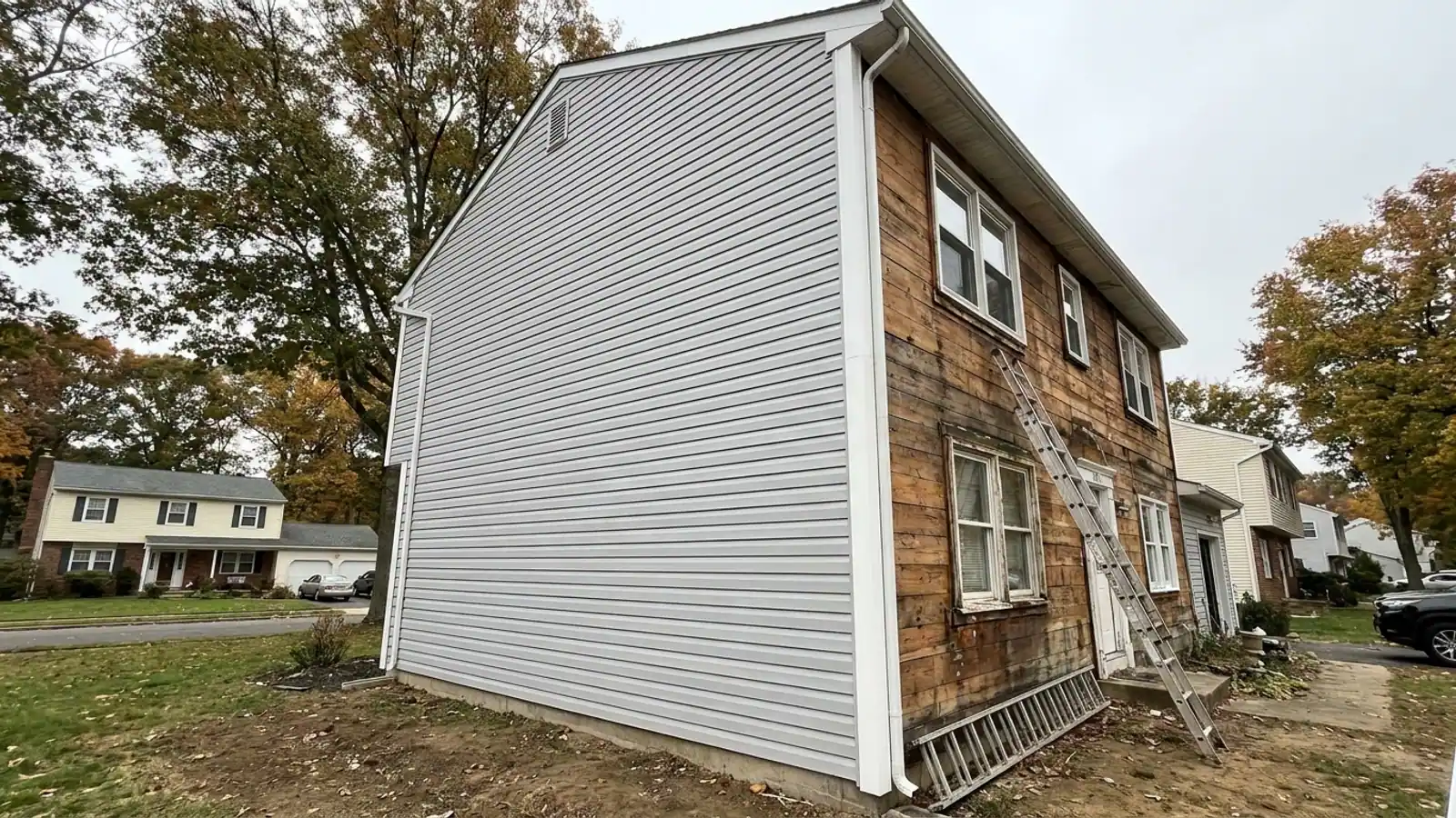 Corner of a two-story house mid-siding-project, new light gray vinyl on one wall and exposed wood sheathing with old trim on the other, ladder set at ground level under overcast sky