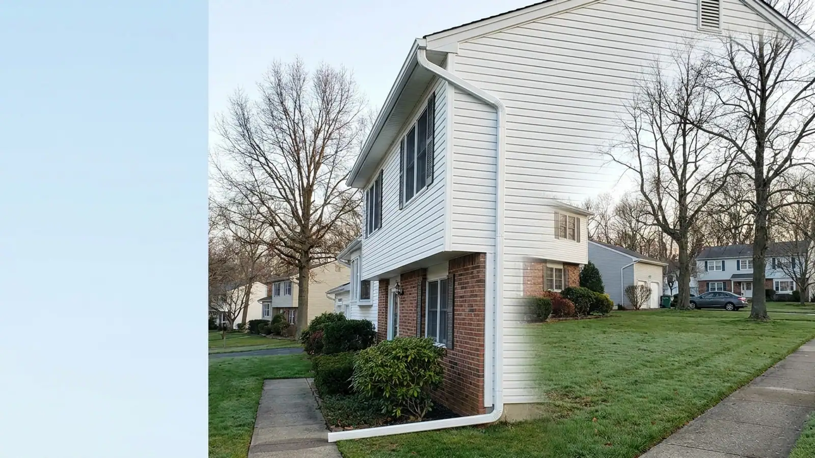 Corner of a two-story colonial home with new white seamless aluminum gutters and a downspout running the full height of the wall, green lawn and foundation shrubs below in morning light
